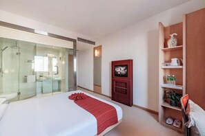 Una habitación de hotel con cama, televisor, estante con artículos de aseo y cabina de ducha de vidrio.