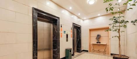 A hallway with marble flooring, two elevators, a trash bin, a reception desk, and a potted plant.