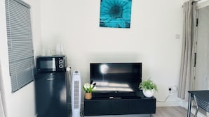 A living room with a flat-screen TV on a black stand, a small kitchenette with a microwave, and a white fan. - Cosy and Comfy Double Room with Private Bathroom, Close to o2 Arena (London)