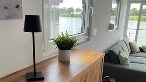 A modern living room with a wooden cabinet, a black floor lamp, a potted plant, a grey sofa, and a view of a lake through large windows.
