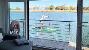 A room with a large window overlooking a lake, a paddleboarder on the water, and a sofa with cushions.