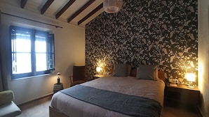 Une chambre avec un grand lit, deux lampes de chevet, une fenêtre avec des stores et un papier peint à motifs floraux.