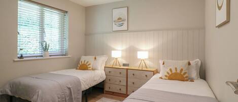 A bedroom with two single beds, a window with blinds, bedside tables with lamps, and a wall-mounted picture.