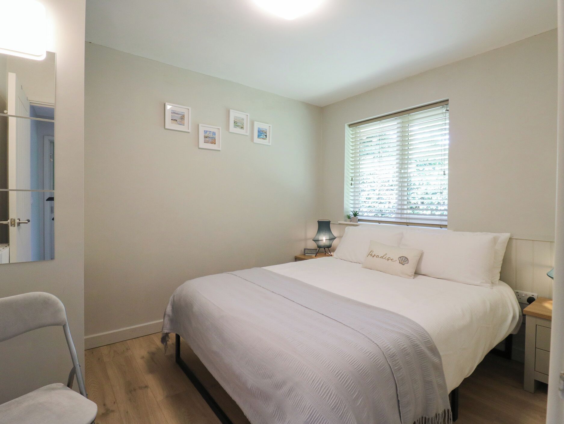 A bedroom with a bed, a chair, a nightstand, a lamp, a window with blinds, and wall-mounted pictures.
