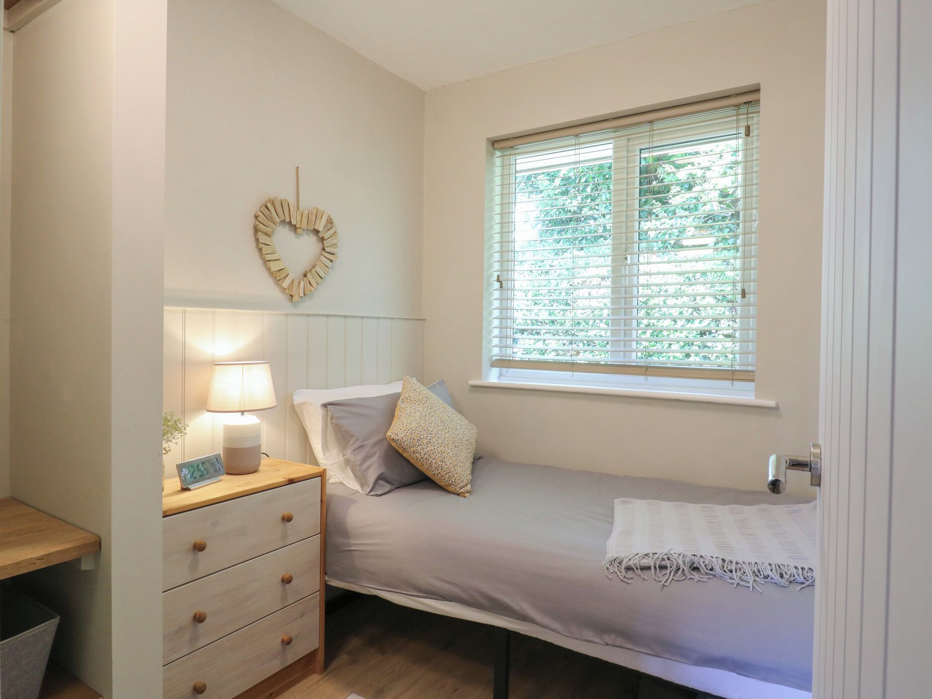 A small, neatly arranged bedroom with a single bed, a bedside table with a lamp, a wall-mounted heart decoration, and a window with blinds.