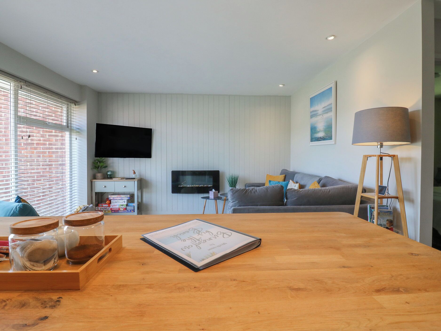 A modern living room with a wooden dining table, grey sofa, and a TV mounted on the wall.