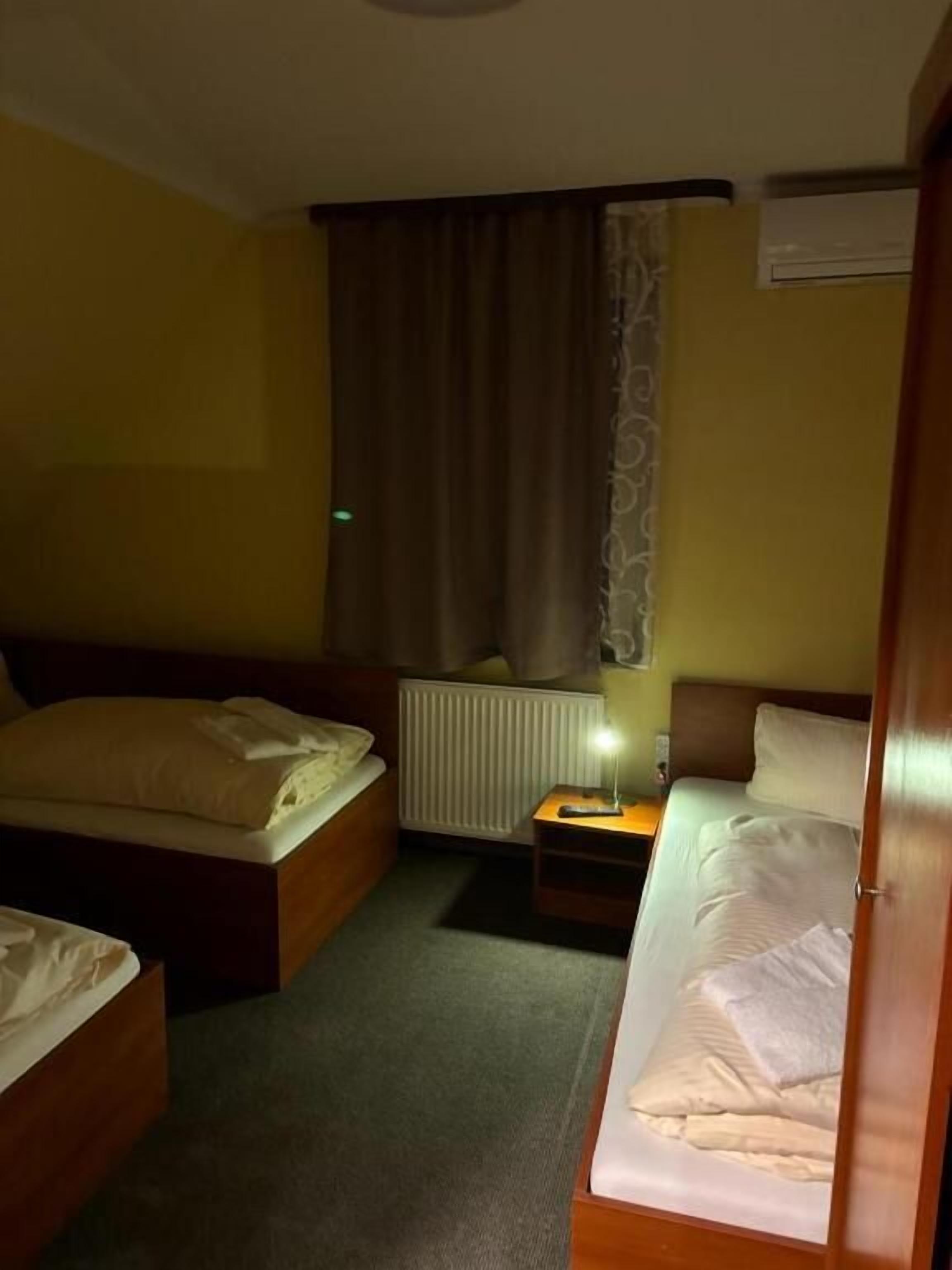 Ein Hotelzimmer mit zwei Einzelbetten, einem Nachttisch mit Lampe, einem Heizkörper und einem Fenster mit Vorhängen.