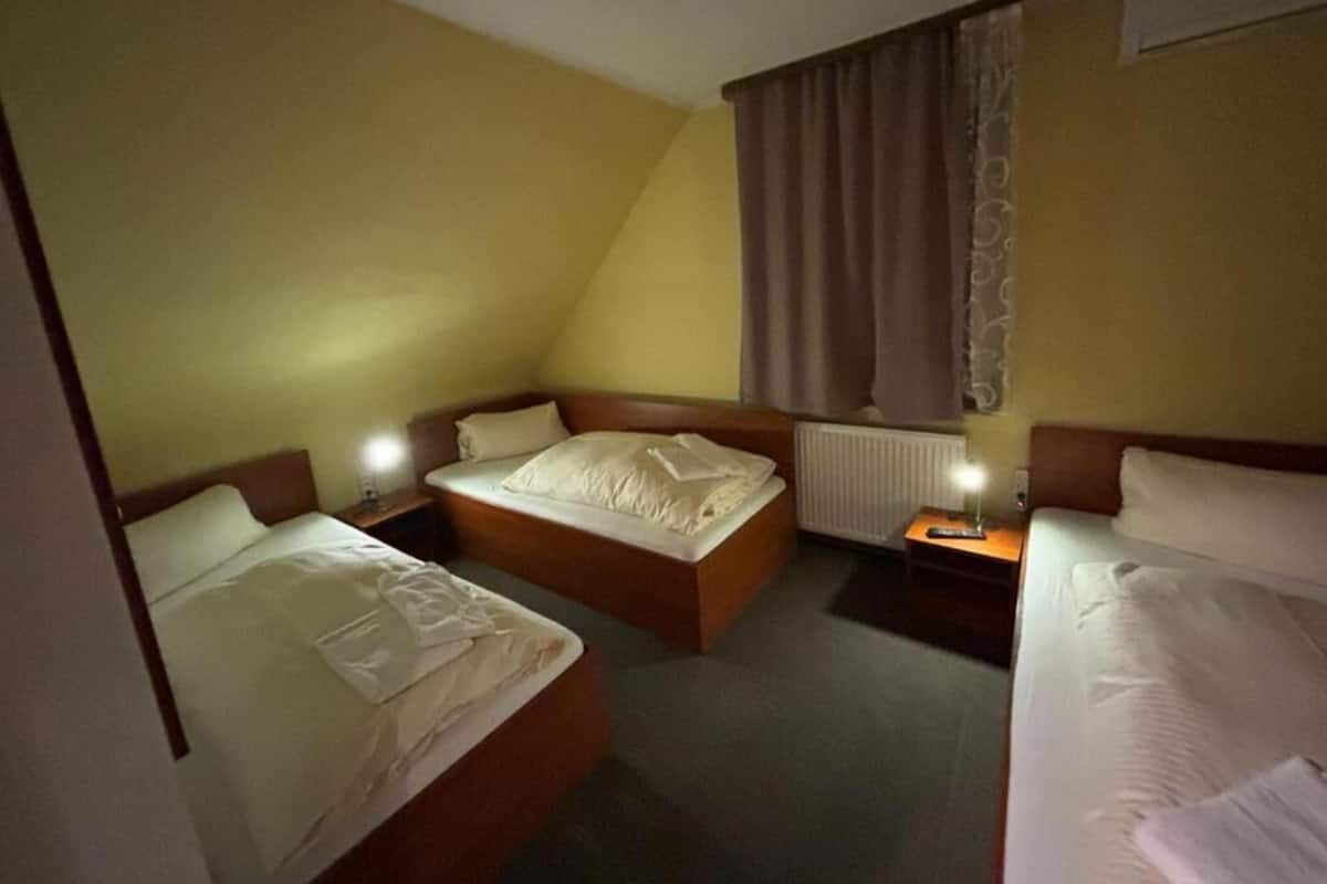 A hotel room with two single beds, a large bed, a nightstand with a lamp, and a window with curtains.