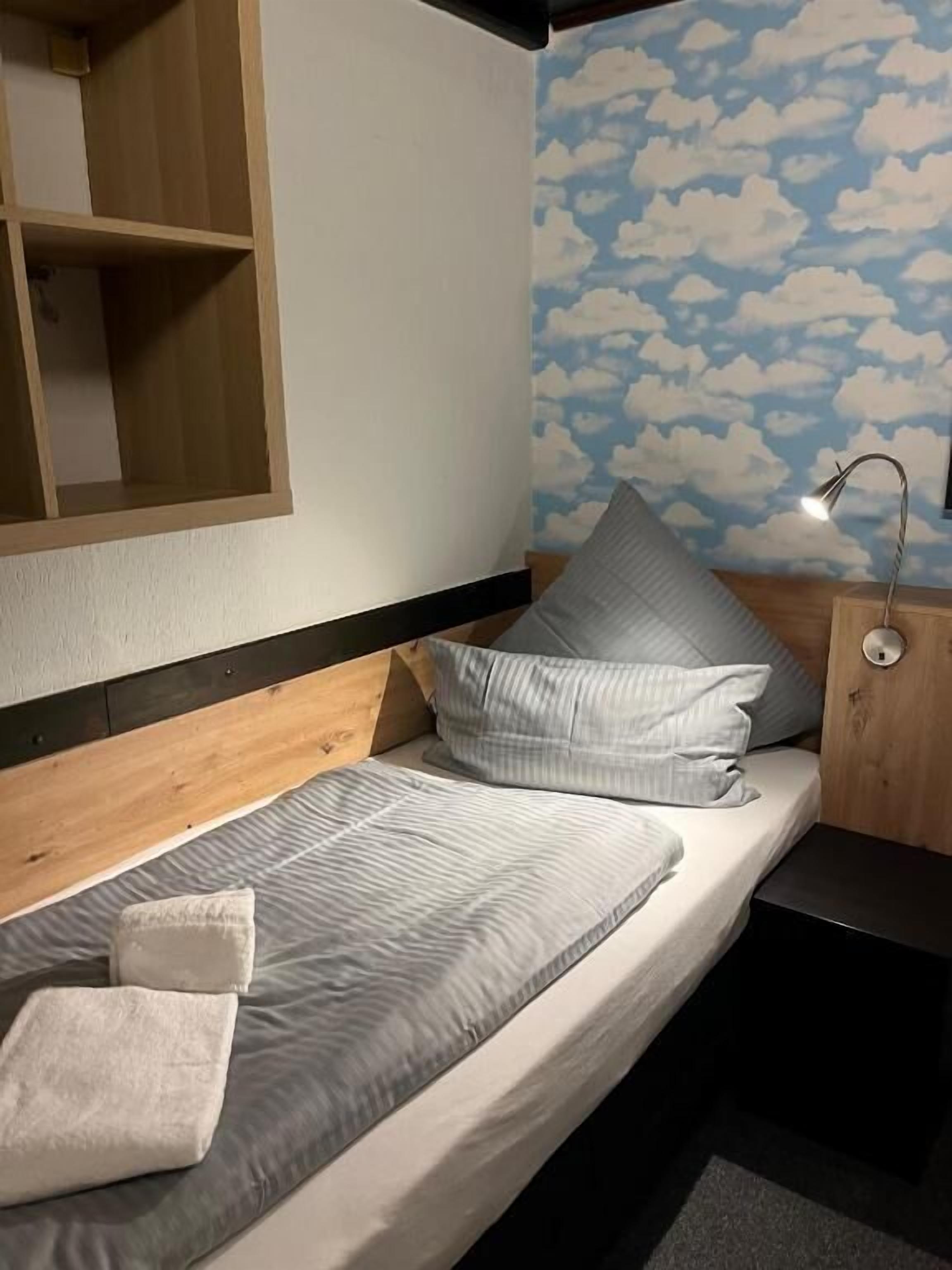 Un llit individual amb llençols i almoades grises, un panell frontal de fusta, una tauleta de nit amb una làmpada i una paret amb un disseny de núvols.