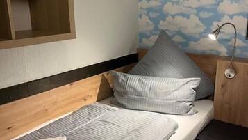 Un llit individual amb llençols i almoades grises, un panell frontal de fusta, una tauleta de nit amb una làmpada i una paret amb un disseny de núvols.
