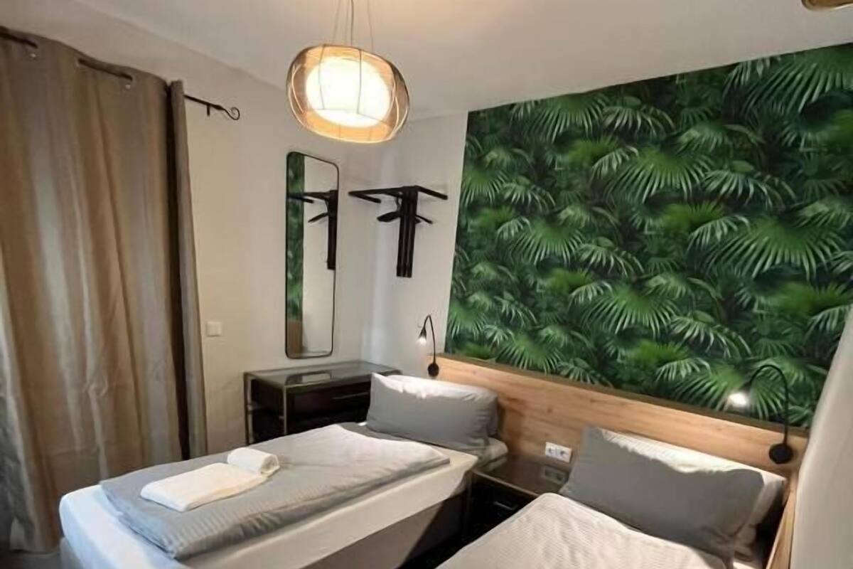 A hotel room with two beds, a large mirror, a ceiling light, and a tropical leaf wallpaper.