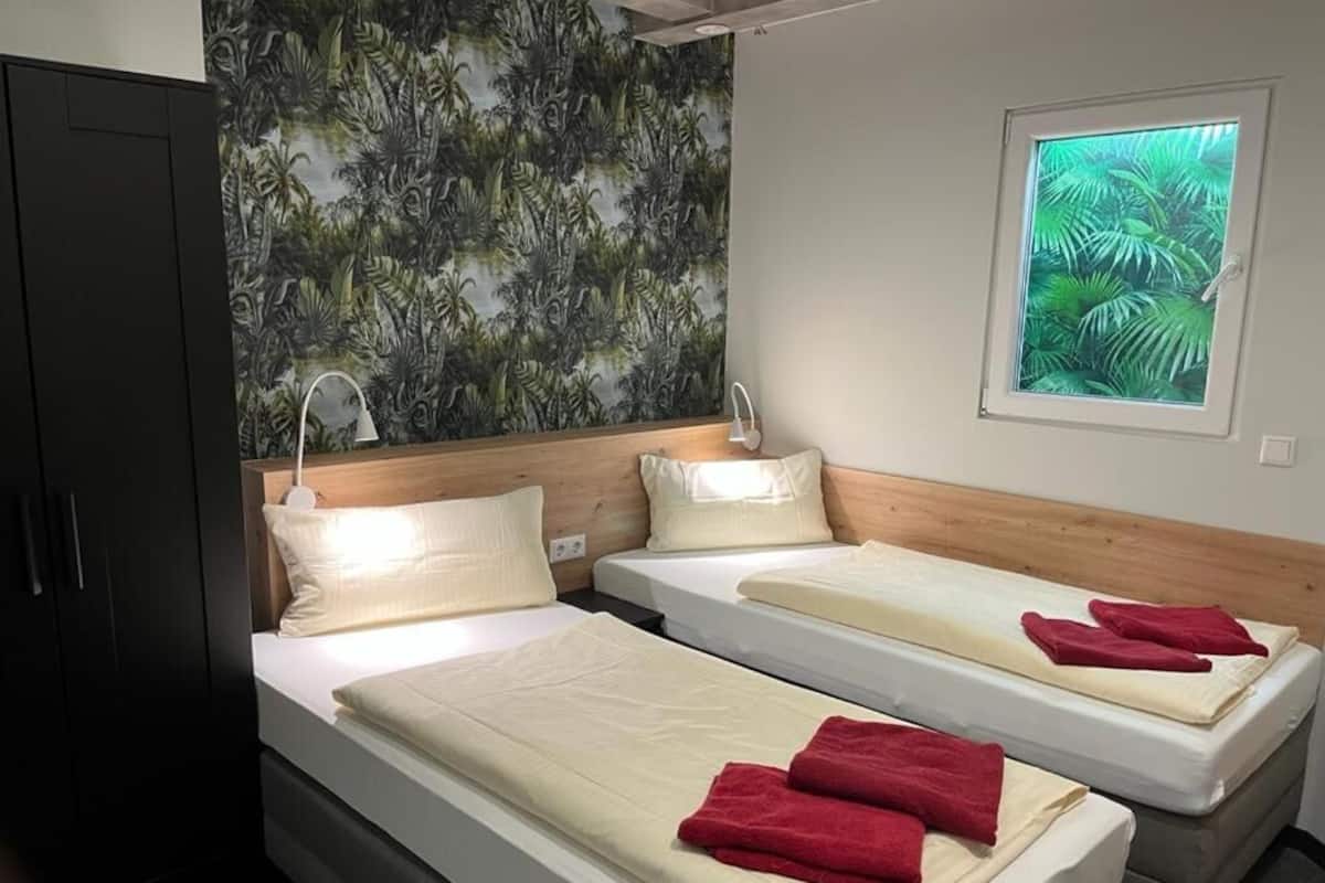 A hotel room with two single beds, a wooden headboard, a large window with a view of greenery, and a dark wardrobe.