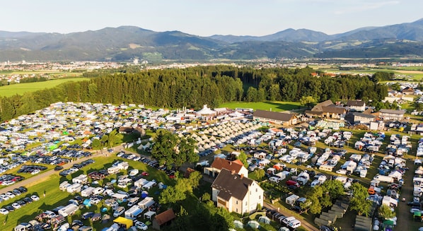 A large outdoor camping area with numerous RVs and cars parked in organized rows, surrounded by green fields and forested hills. - RacingTours RaceCamp (Spielberg)