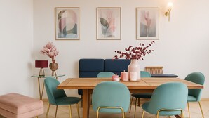 A dining area with a wooden table, teal chairs with gold legs, a pink ottoman, and a vase with red flowers. There are three framed abstract paintings on the wall and a wall-mounted light fixture.