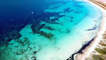Vue aérienne d’une zone côtière avec des eaux turquoise limpides, une plage de sable et un promontoire rocheux situé au loin.