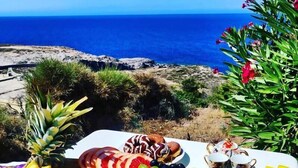 A table with a variety of fruits, pastries, and drinks, set against a scenic view of the ocean.