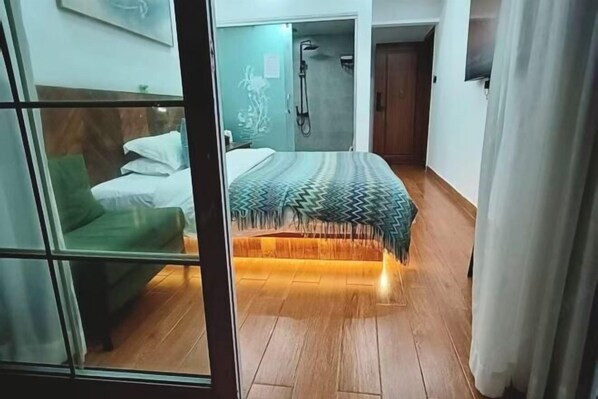 A hotel room with a bed, a green armchair, a shower area, and a wooden floor.