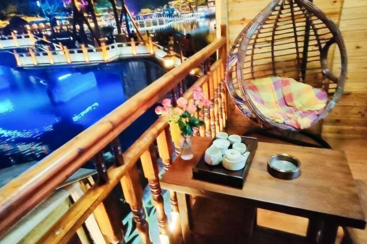 A wooden balcony with a hanging chair, a small table with a vase of flowers, and a view of boats on water.