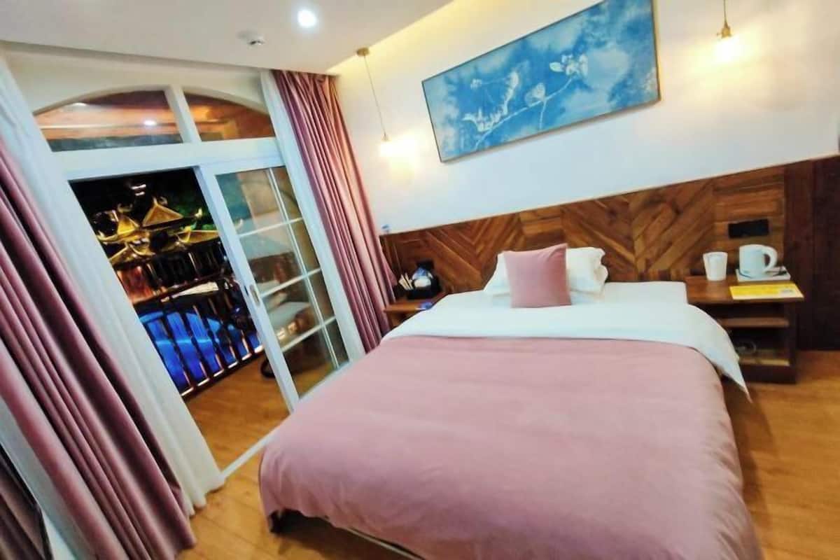 A hotel room with a bed, a nightstand, a painting, and a view of a building through a glass door.