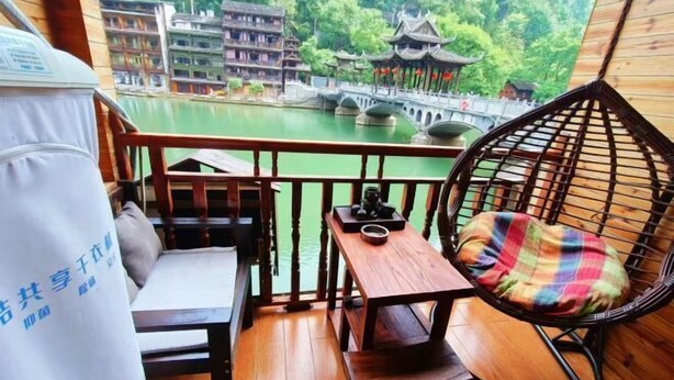 A balcony with a wooden table, a chair with a plaid cushion, and a view of traditional buildings and a bridge over a body of water. - Fengyuanju Inn (Fenghuang County)