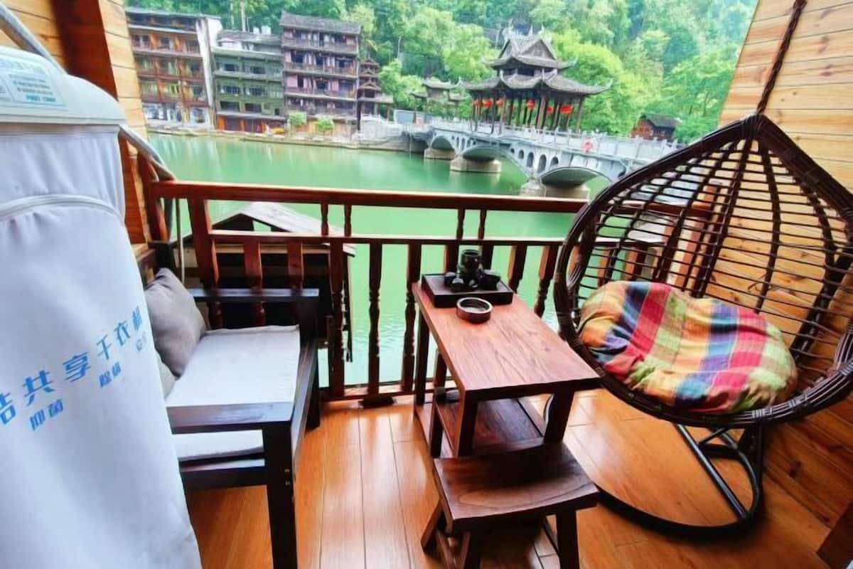 A balcony with a wooden table, a chair with a plaid cushion, and a view of traditional buildings and a bridge over a body of water.