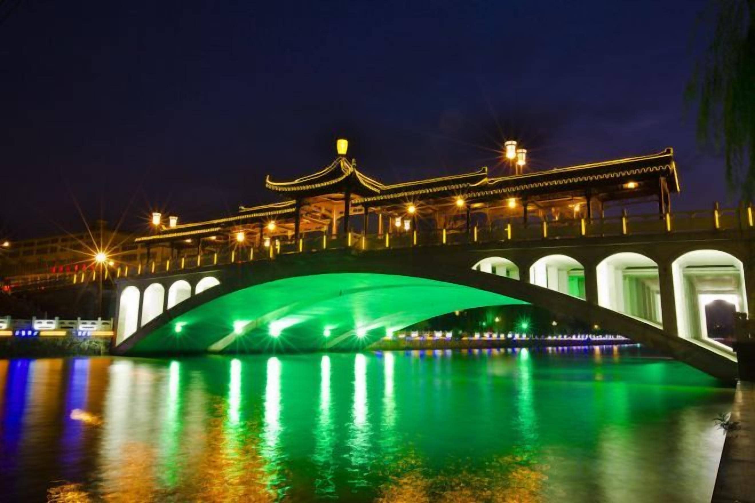 Un pont avec une arche verte qui enjambe l’eau, dont les lumières se reflètent à la surface, et un bâtiment à l’arrière-plan, doté d’une architecture traditionnelle.