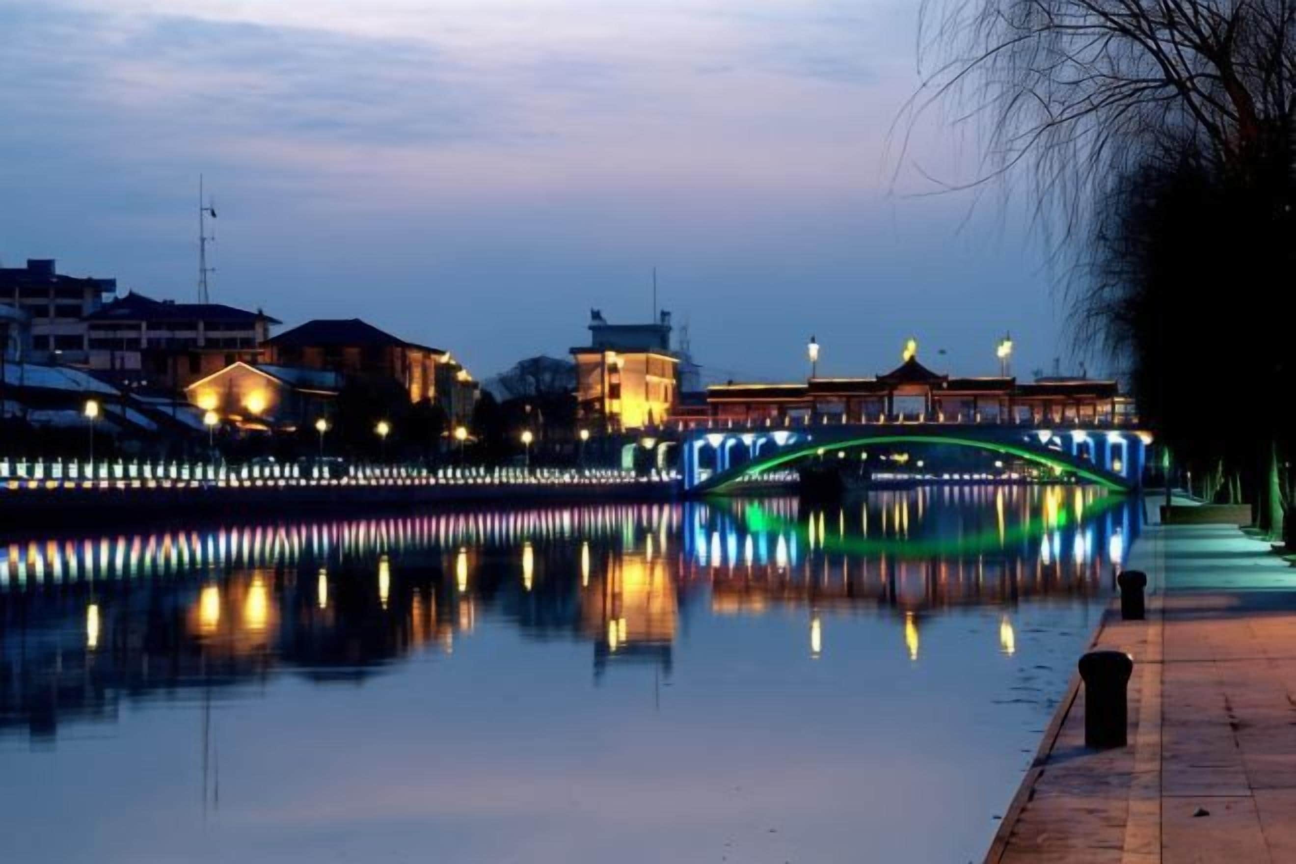 A calm river with a bridge reflecting lights, a building with a traditional roof, and a clear sky.