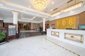 A hotel lobby with a reception desk, a chandelier, and a staircase leading to the upper floor.