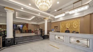 Un vestíbulo de hotel con piso de mármol, una gran araña y un mostrador de recepción con dos empleados, además de una escalera que conduce a otra zona.