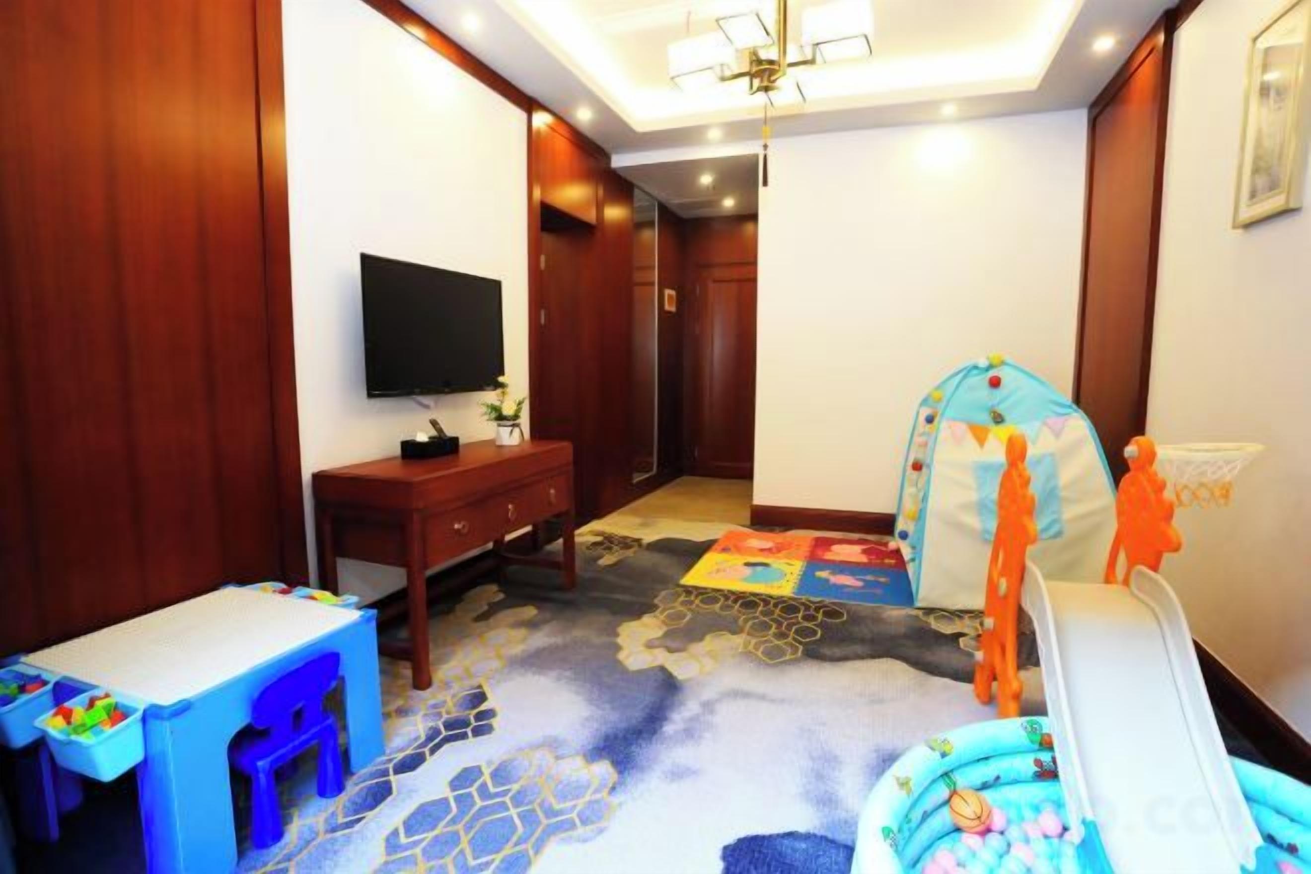 A hotel room with a television, a desk, a chair, a bed with a colorful blanket, and a play area with toys.