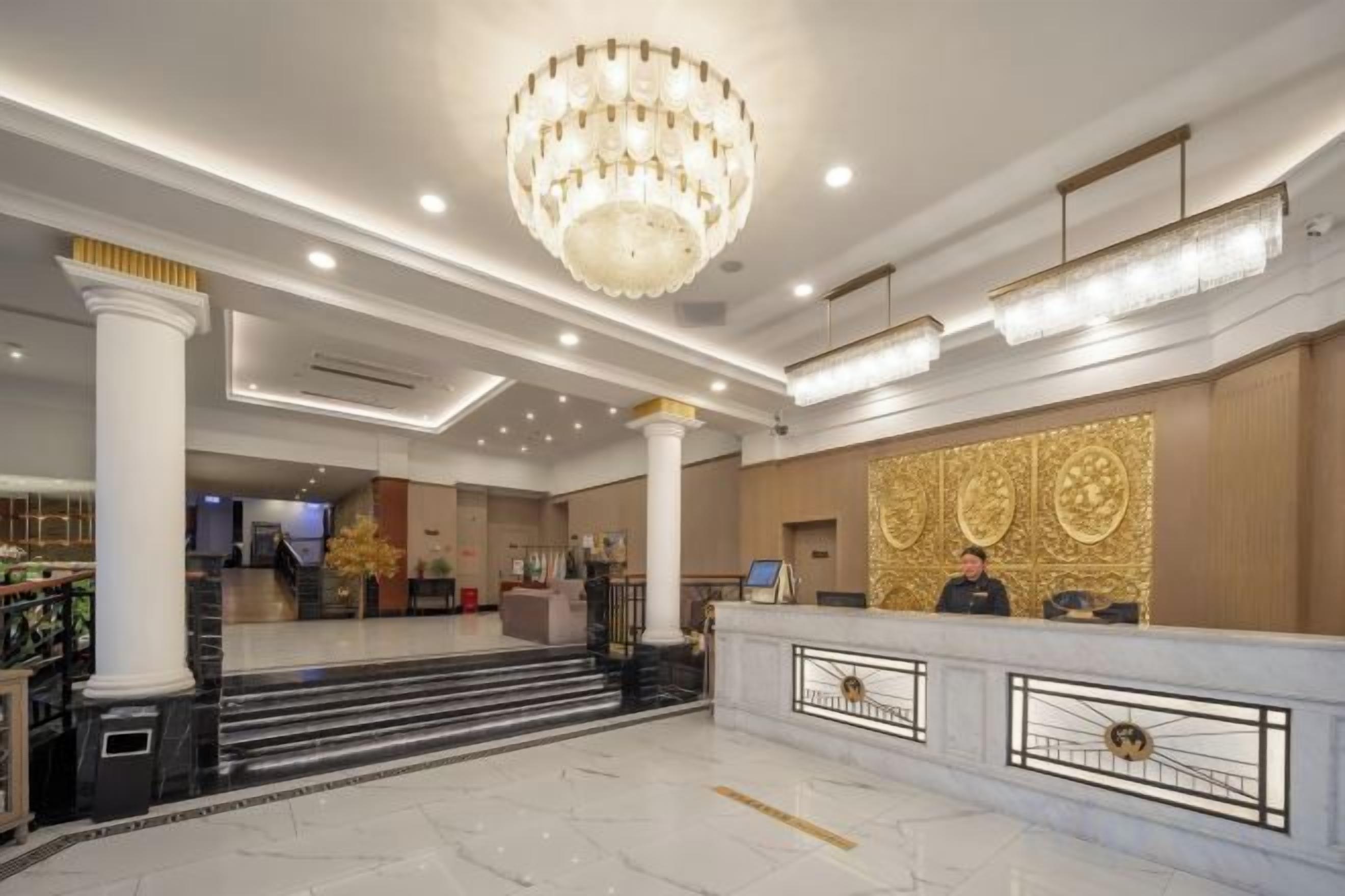A hotel lobby with marble flooring, a grand chandelier, a reception desk, and a staircase leading to the upper floor.