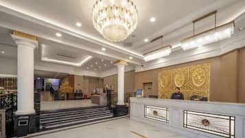 A hotel lobby with marble flooring, a grand chandelier, a reception desk, and a staircase leading to the upper floor.