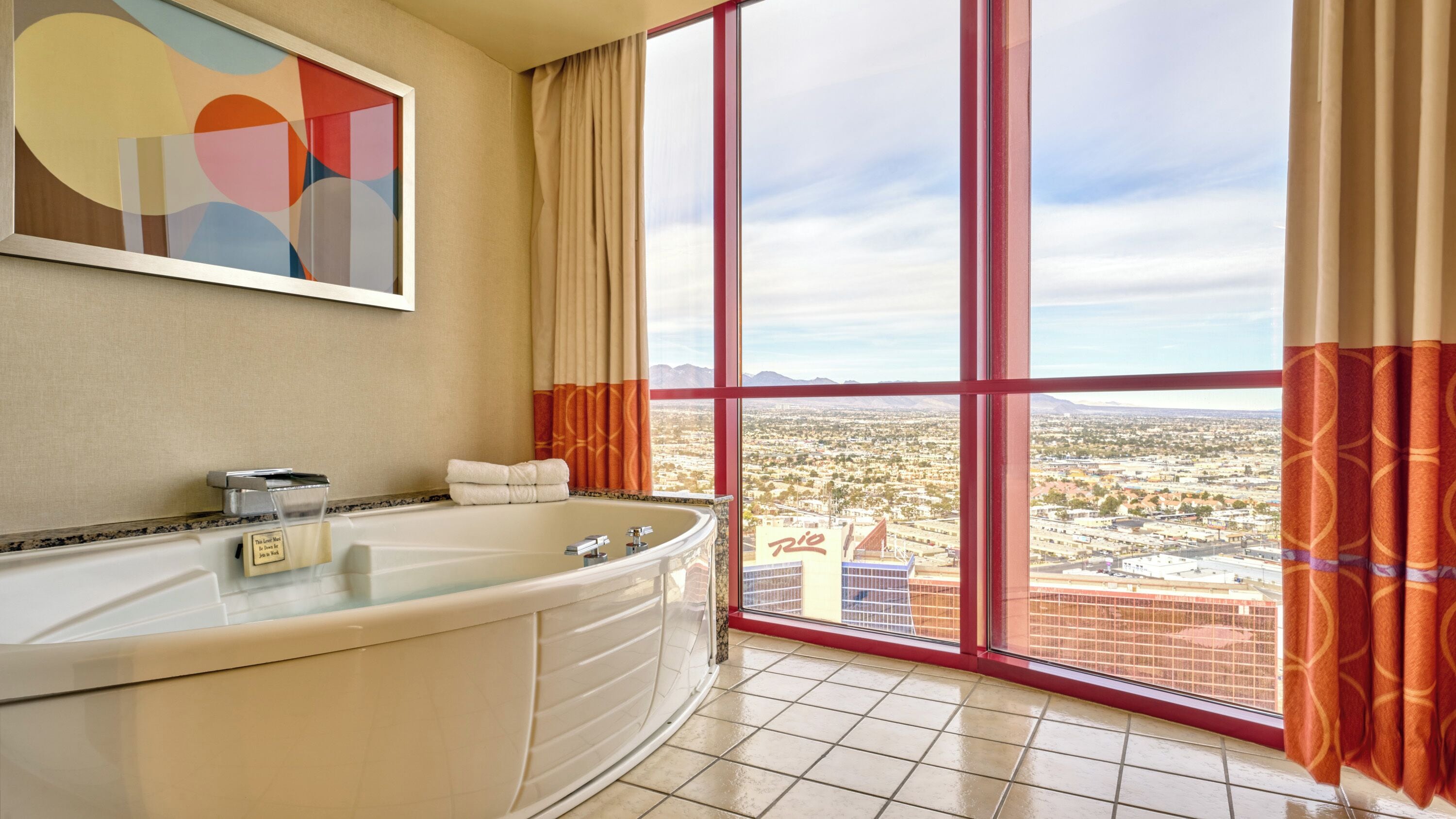 A bathroom with a large bathtub, a mirror, and a view of a cityscape through large windows.