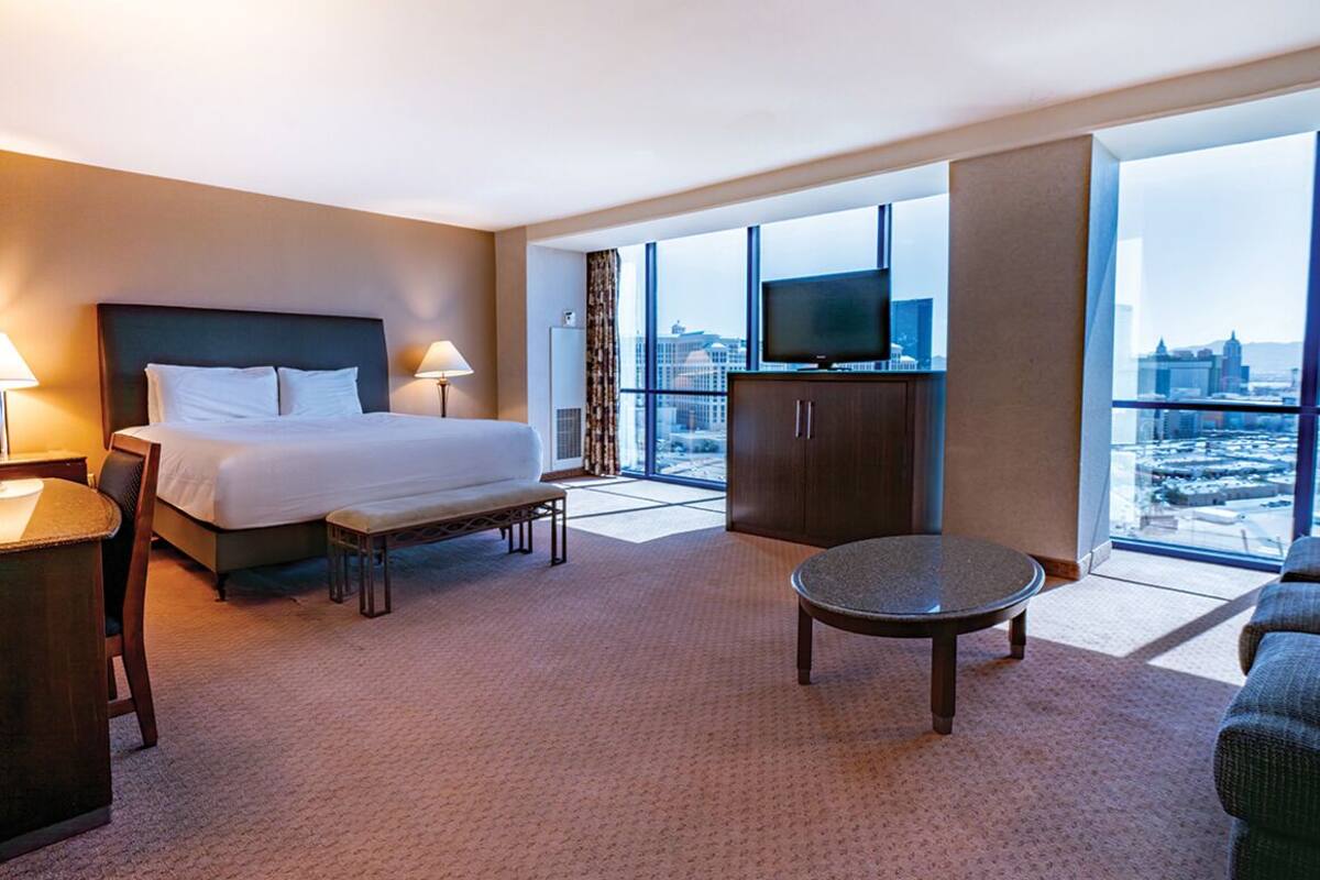 A hotel room with a bed, a desk, a chair, a round table, a television, and a large window with a city view.