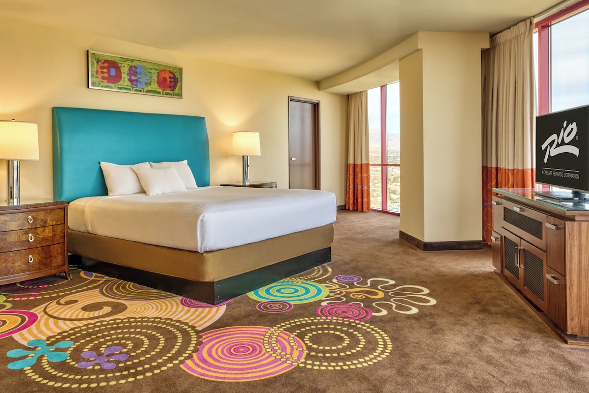 A hotel room with a bed, two bedside lamps, a television, a desk with a computer, and a colorful carpet.