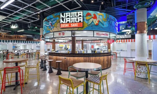 Una barra de sushi y sake con un mural colorido, rodeada de mesas y sillas en un espacio interior amplio.