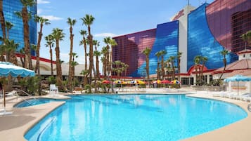 A large indoor swimming pool with lounge chairs and umbrellas, surrounded by palm trees and modern buildings with reflective glass facades.