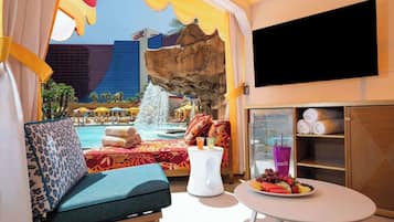 Een hotelkamer met uitzicht op een zwembad en een waterfontein, een bed met een kleurrijk sprei, een klein tafeltje met fruit en drankjes, en een aan de muur bevestigde televisie.