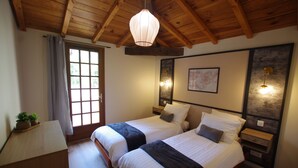 Una habitación con dos camas, vigas de madera en el techo, una ventana con cortinas, lámparas de noche y una pequeña mesa con una planta.