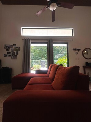 Una habitación con un sofá rojo, un ventilador de techo, una ventana con cortinas y una pared con cuadros.