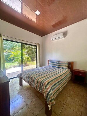 Un dormitorio con cama, unidad de aire acondicionado, mesita de noche de madera y puerta corrediza de vidrio que da a un jardín.