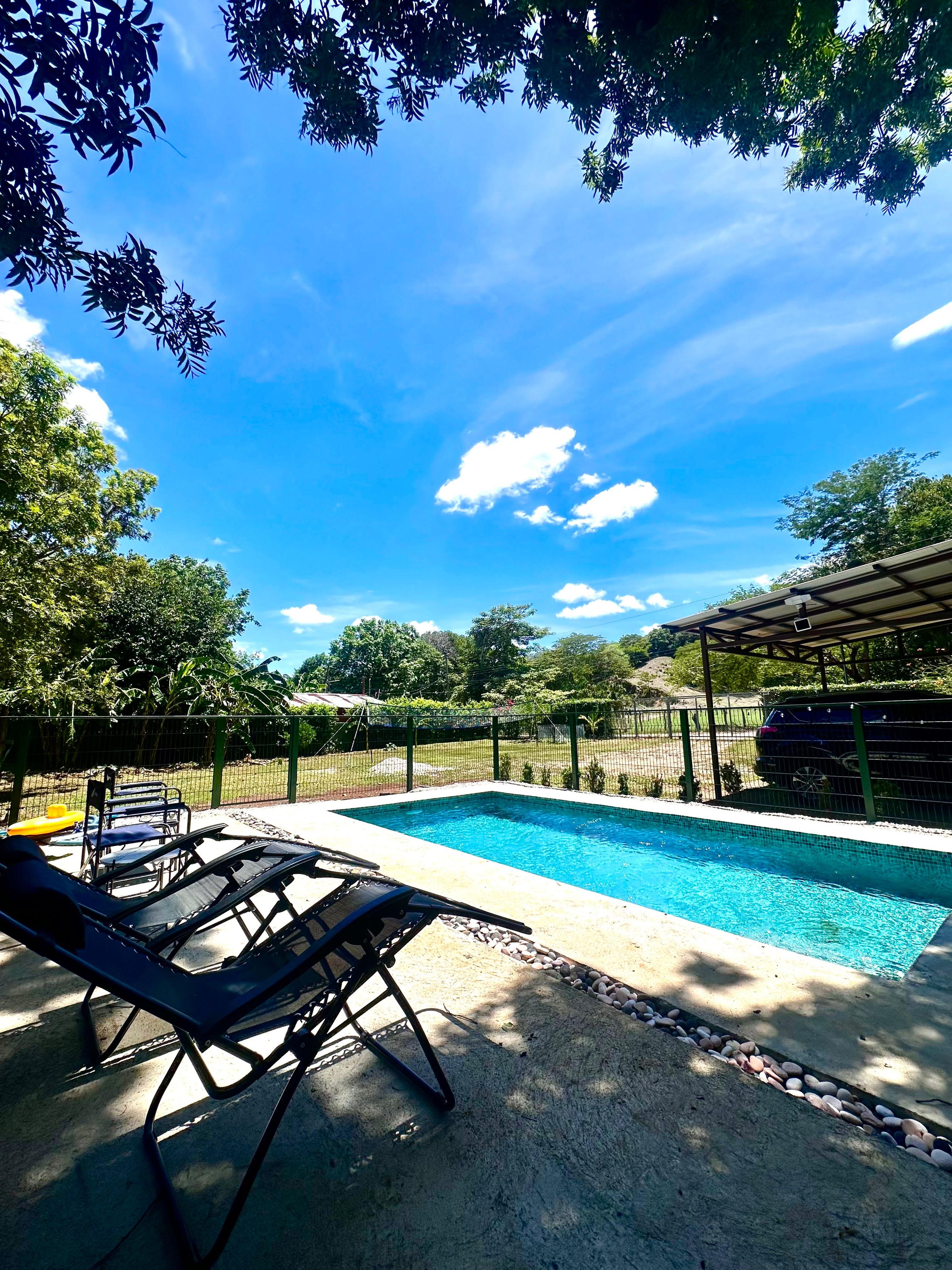 A swimming pool with lounge chairs and a covered area, surrounded by a fence and trees.