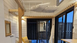 A cozy room with a wooden floor, a grey sofa with a yellow pillow, a small round table, and a large window with a view of the outside. - Tiny House with private Spa (Boigny-sur-Bionne)