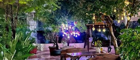 Un patio al aire libre con una mesa y sillas de madera, rodeado de plantas en macetas y árboles.