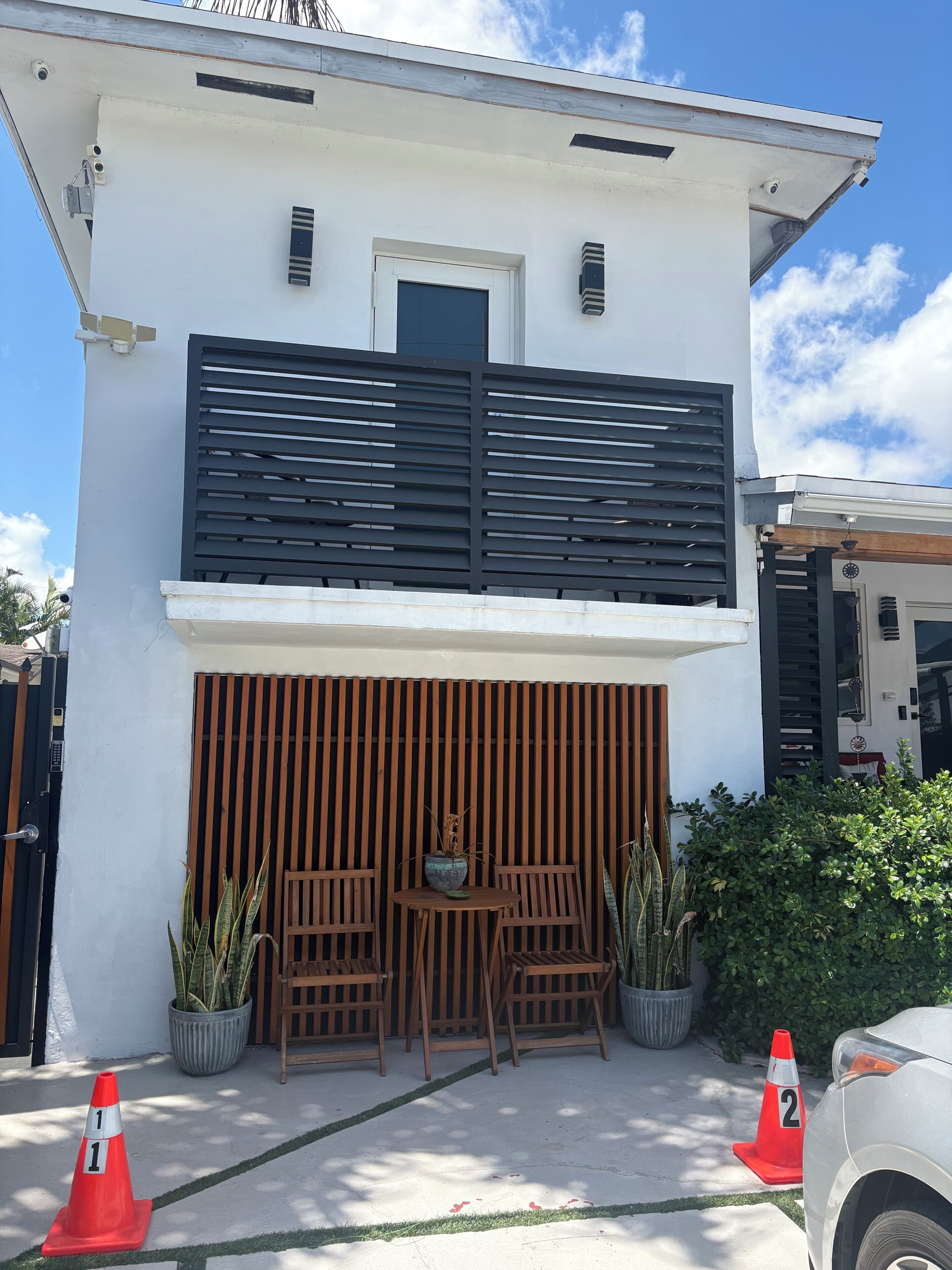 A two-story building with a white facade, a black slatted balcony, and a wooden fence at the entrance. There are two potted plants and a table with chairs outside.