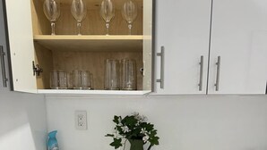 Une cuisine avec un plan de travail blanc, deux tabourets de bar noirs, un vase vert avec des fleurs et une étagère contenant des verres à vin et des verres à cocktail.
