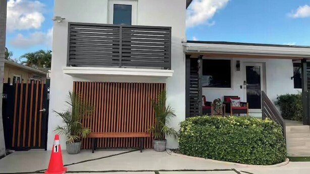 Une maison moderne à deux étages, dotée d’un toit plat, d’une clôture en bois et d’un petit porche aménagé avec du mobilier d’extérieur.
