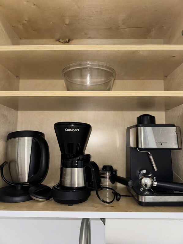 A kitchenette with a coffee maker, a kettle, and a water dispenser on a wooden shelf.
