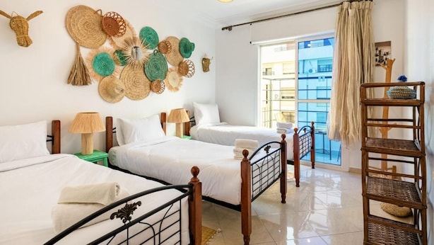 A hotel room with two beds, a wicker shelf, and a view of the outside.