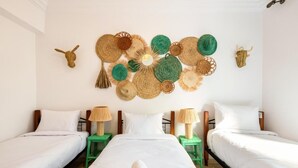 A hotel room with two single beds, a large wall hanging of woven items, and two small tables with lamps.